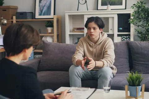Un adolescent en chandail à capuchon, assis sur un canapé, parle à une intervenante qui l’écoute.
